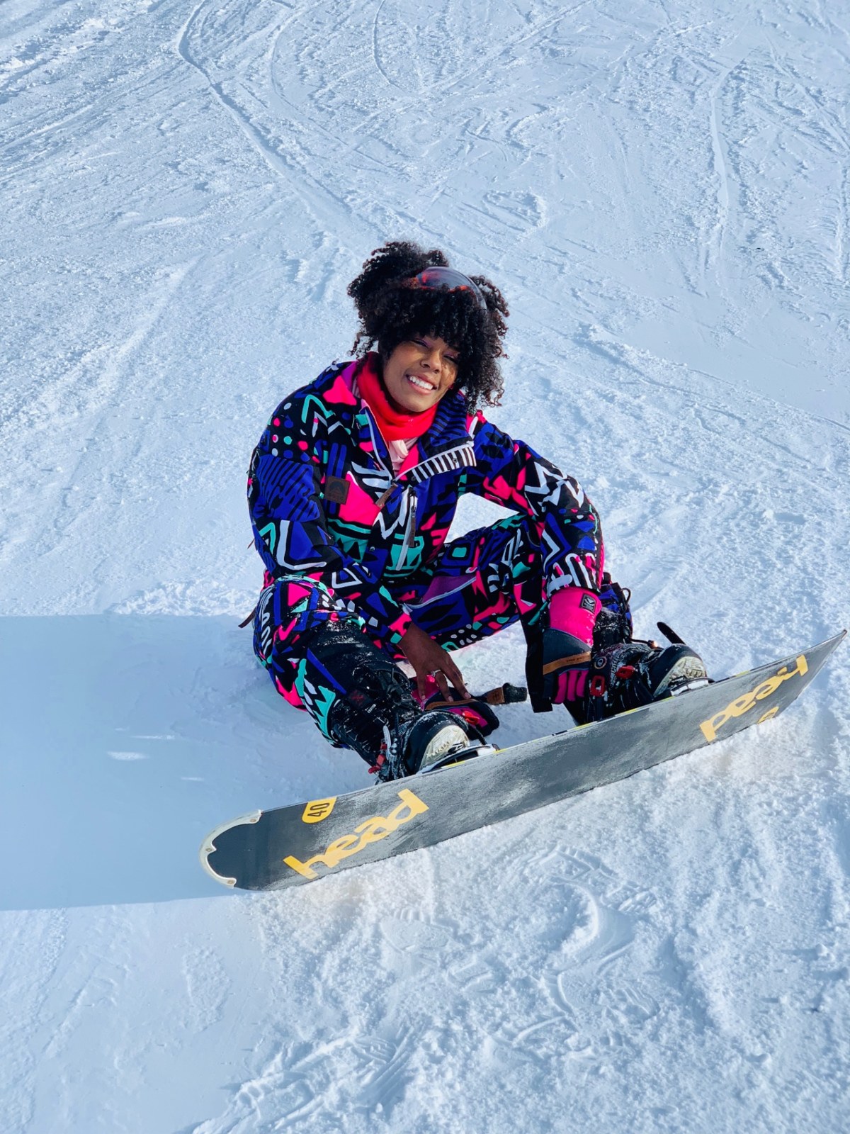 Sojourn Wallace sitting in the snow in a colorful patterned snowboard suit